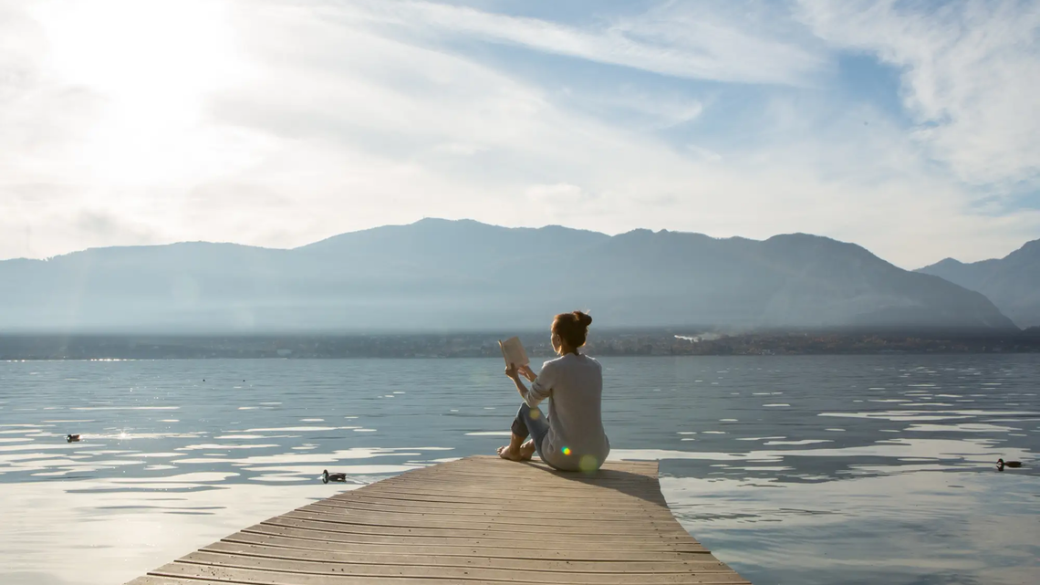 Frau liest ein Buch auf einem Steg am See | © GettyImages/	swissmediavision