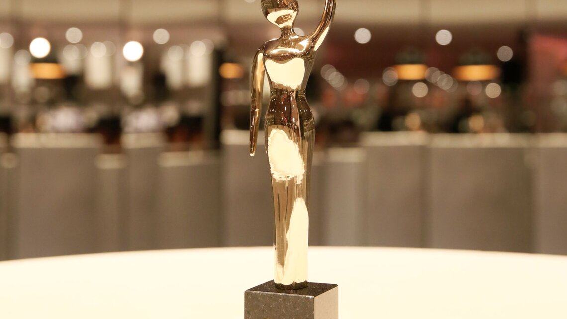 Eine glänzende, goldene Trophäe in menschlicher Form steht auf einem schwarzen Sockel vor einem verschwommenen, hell erleuchteten Hintergrund. | © GOLDENE BILD der FRAU